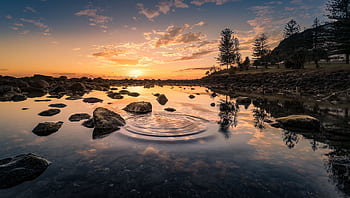 Peaceful river scene at sunrise with rippling water, rocks, and trees. HD wallpaper