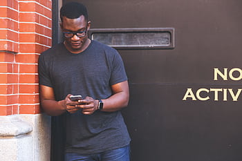 African American man using smart while leaning against a brick wall, wearing casual attire. HD wallpaper