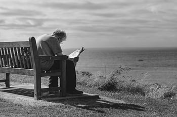 A contemplative scene of an elderly man reading a newspaper on a seaside bench with cloudy skies. HD wallpaper