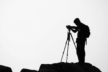 A grapher with a backpack uses a tripod camera on a cliff's edge during a cloudy day. HD wallpaper