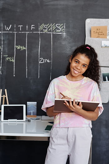 Teen girl smiles while studying indoors, holding a notebook in front of a blackboard. HD phone wallpaper
