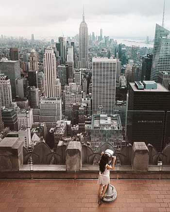 A woman using binoculars to view the iconic New York City skyline from a rooftop. HD phone wallpaper