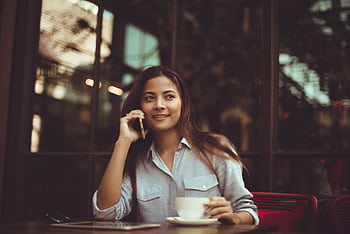 Young woman in casual shirt talking on and holding coffee at an outdoor café table. HD wallpaper