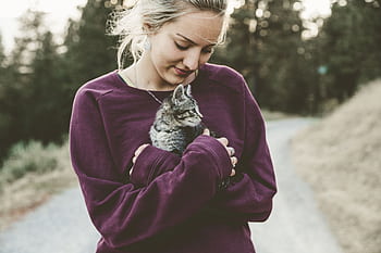 Young woman in a purple sweater smiling down at a kitten she's holding outdoors on a pathway. HD wallpaper