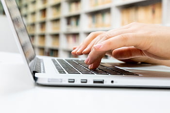 Close-up of hands typing on a laptop keyboard in a library setting. HD wallpaper