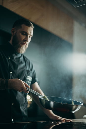 A focused chef prepares a meal using a frying pan in an indoor kitchen setting. HD phone wallpaper