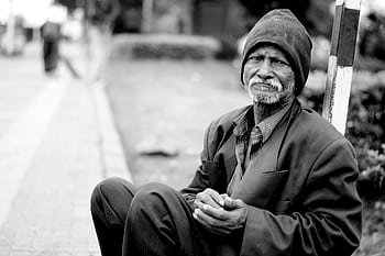 A black and white portrait of an elderly man sitting on the streets, expressing hardship and resilience. HD wallpaper