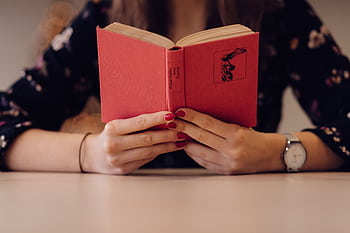 Close-up of a woman reading a red book, showcasing her painted nails and wristwatch in an indoor setting. HD wallpaper