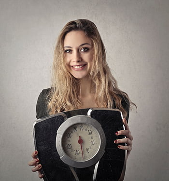 Young woman with blond hair smiling, holding a vintage bathroom scale indoors. HD phone wallpaper