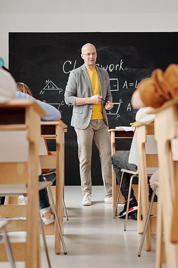 Bald teacher explaining geometry on blackboard to attentive students in classroom. HD phone wallpaper
