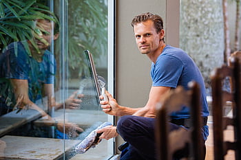 A man in a blue t-shirt cleaning large glass pane with a squeegee indoors. HD wallpaper