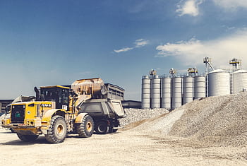 Yellow loader and truck operating at a gravel yard with silos in the background on a sunny day. HD wallpaper