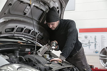A mechanic closely inspects a car engine in a garage using a flashlight, ensuring proper maintenance. HD wallpaper