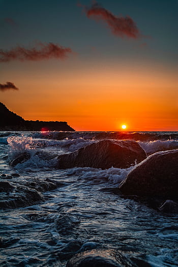 Dramatic sunset over ocean waves crashing onto rocky shore. Perfect for serene and nature-themed content. HD phone wallpaper