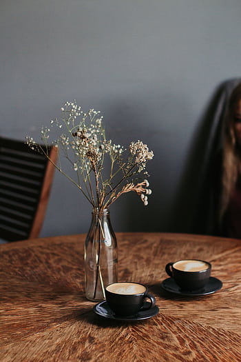 Two cappuccino cups with frothy art on a wooden table, accompanied by a delicate flower vase. HD phone wallpaper