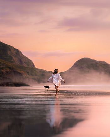 A serene beach scene with a woman and her dog enjoying a run at sunset. HD phone wallpaper