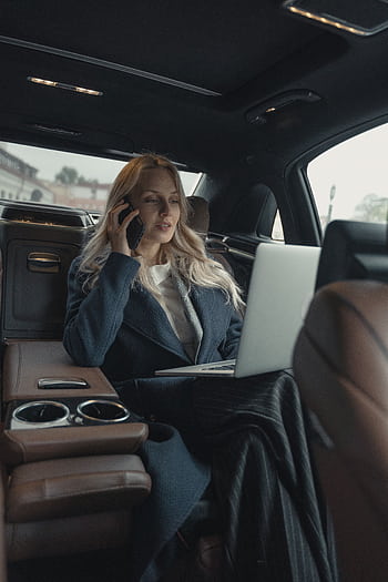 Confident businesswoman making a call and working on a laptop in a luxury car interior. HD phone wallpaper