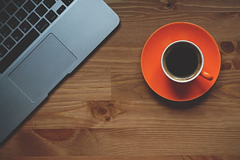 Top-down shot of a coffee cup on an orange saucer next to a laptop on a wooden desk surface. HD wallpaper