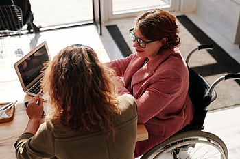 Two women working on laptops, one using a wheelchair, collaborating in a modern café. HD wallpaper