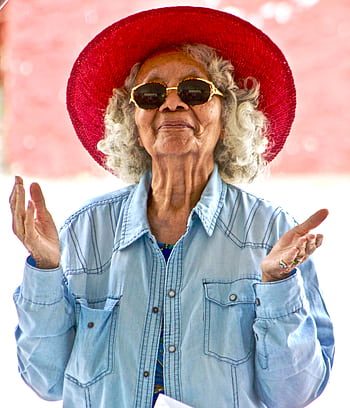 Joyful senior woman wearing a vibrant red hat and sunglasses, expressing happiness outdoors. HD phone wallpaper