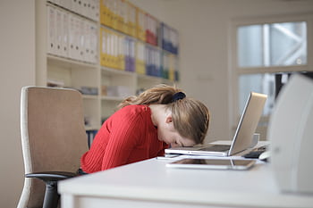 A tired woman in a red sweater leans her head on a desk with a laptop, symbolizing workplace fatigue. HD wallpaper