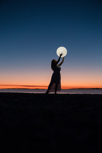 A serene scene of a woman silhouetted against a sunset sky, reaching towards the full moon. HD phone wallpaper