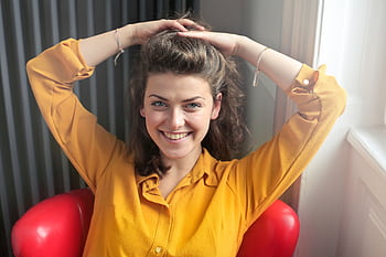 Cheerful woman sitting indoors, smiling brightly with hands behind head, wearing a yellow shirt. HD wallpaper