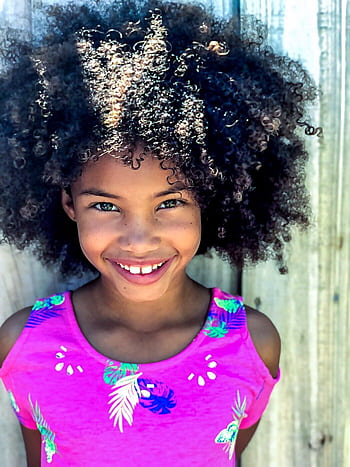 A joyful young girl showcases her beautiful curly Afro hair and bright smile in an outdoor portrait against a wooden backdrop. HD phone wallpaper