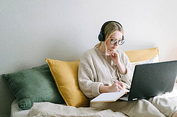 A focused woman in glasses and heads works on a laptop from a cozy bed. HD wallpaper