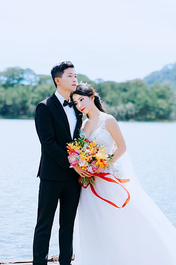 Elegant bride and groom posing by a lake with colorful bouquet. HD phone wallpaper