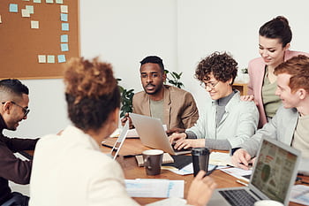 Multicultural team engaged in a collaborative office meeting, discussing ideas around a table with laptops. HD wallpaper