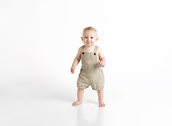 Adorable baby in beige overalls standing against a white background, showcasing innocence and joy. HD wallpaper
