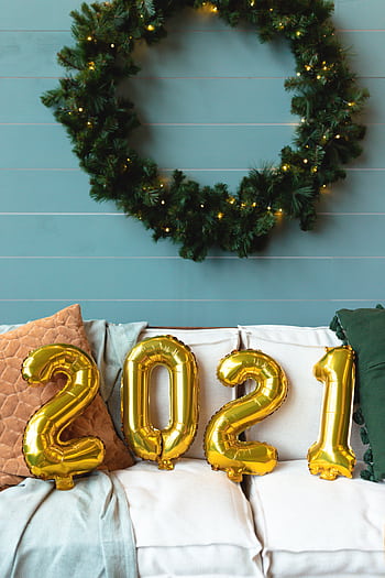 A cozy Christmas interior decor with golden '2021' balloons on a sofa beneath a lit wreath. HD phone wallpaper
