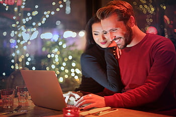 A couple using a laptop at a cozy restaurant with festive bokeh lights, enjoying online shopping together. HD wallpaper