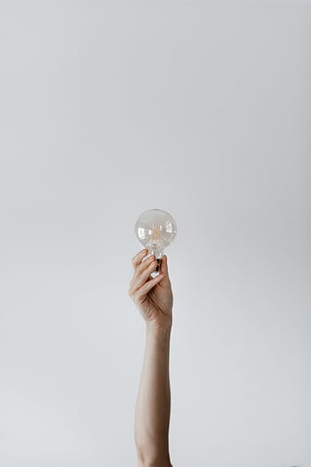 A hand raises a light bulb against a soft gray background, symbolizing creativity and ideas. HD phone wallpaper