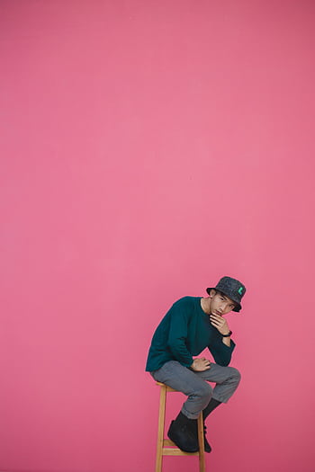 Man in fashionable attire posing on a stool with a pink background, exuding style and confidence. HD phone wallpaper
