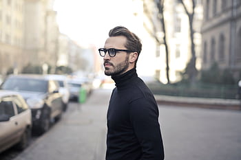 A fashionable man in a black top and glasses poses in an urban street with parked cars. HD wallpaper