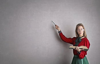 Female teacher pointing at a blank wall, holding a book, creating copyspace for educational content. HD wallpaper