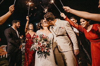 A joyful couple kisses under sparklers, surrounded by guests in a wedding celebration. HD wallpaper