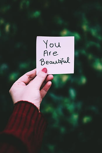 A close-up of a hand holding a 'You Are Beautiful' note against a blurred green background. HD phone wallpaper