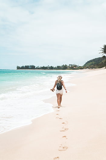 A woman with a backpack walks barefoot along a pristine tropical beach, leaving footprints in the sand. HD phone wallpaper