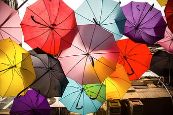 A dynamic array of colorful umbrellas creates a vibrant canopy in this lively outdoor street scene. HD wallpaper