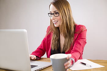 A smiling young businesswoman in a red jacket works on her laptop while holding a coffee cup. HD wallpaper