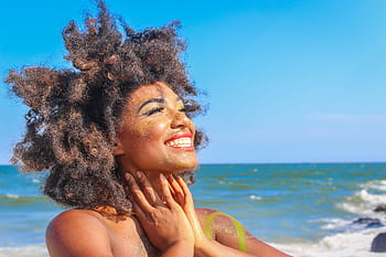 A joyful woman with afro hair smiling brightly on a sunny beach day. HD wallpaper