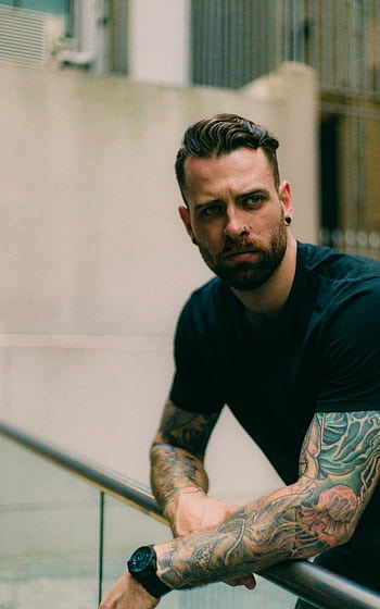 Portrait of a stylish, tattooed man leaning on a railing with a contemplative expression. HD phone wallpaper