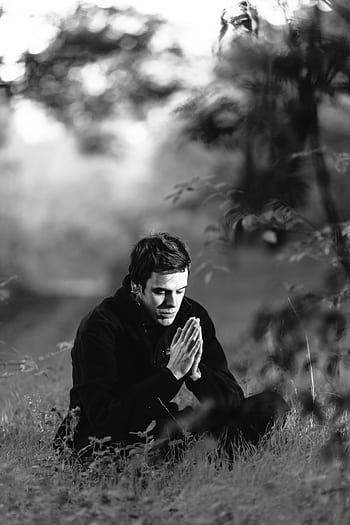 A man sitting in a grassy field, meditating with hands in prayer position, captured in black and white. HD phone wallpaper