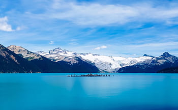 A stunning view of a serene mountain lake with snow-capped peaks under a clear blue sky. HD wallpaper