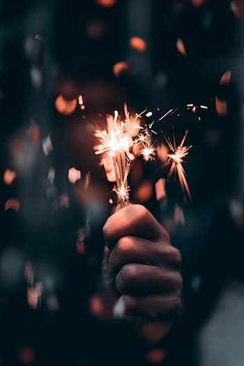 A close-up of a hand holding a glowing sparkler, creating a festive night scene. HD phone wallpaper