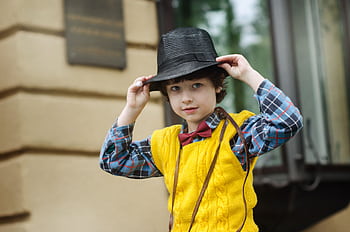 A stylish young boy wearing a yellow sweater, plaid shirt, and black hat poses playfully outdoors. HD wallpaper