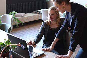 Two professional women discuss a project using a laptop in a modern office environment. HD wallpaper